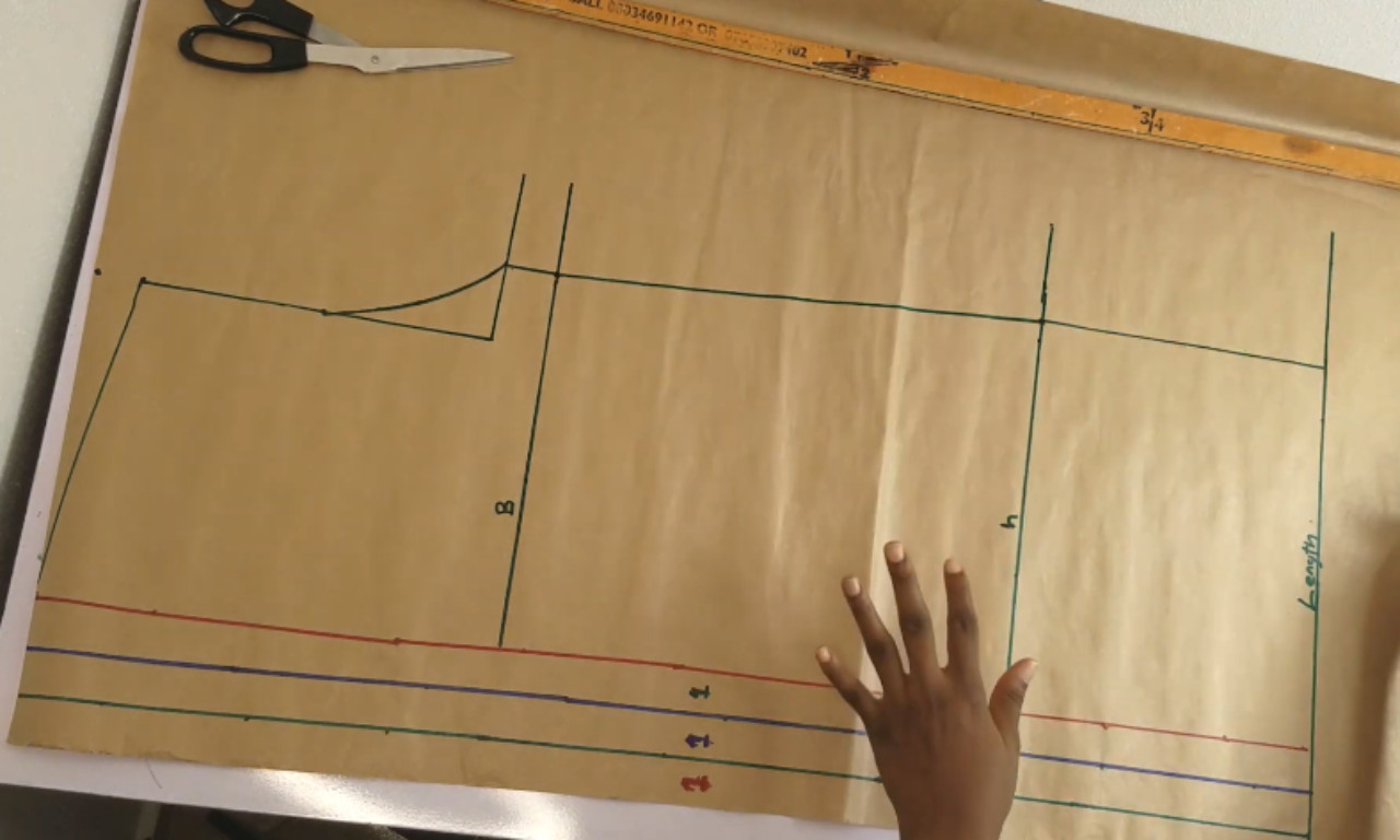 A hand placed on a large brown paper dressmaking pattern with neckline, shoulder, and body outlines drawn in green, alongside measurement lines in red and blue; scissors and a ruler are nearby.