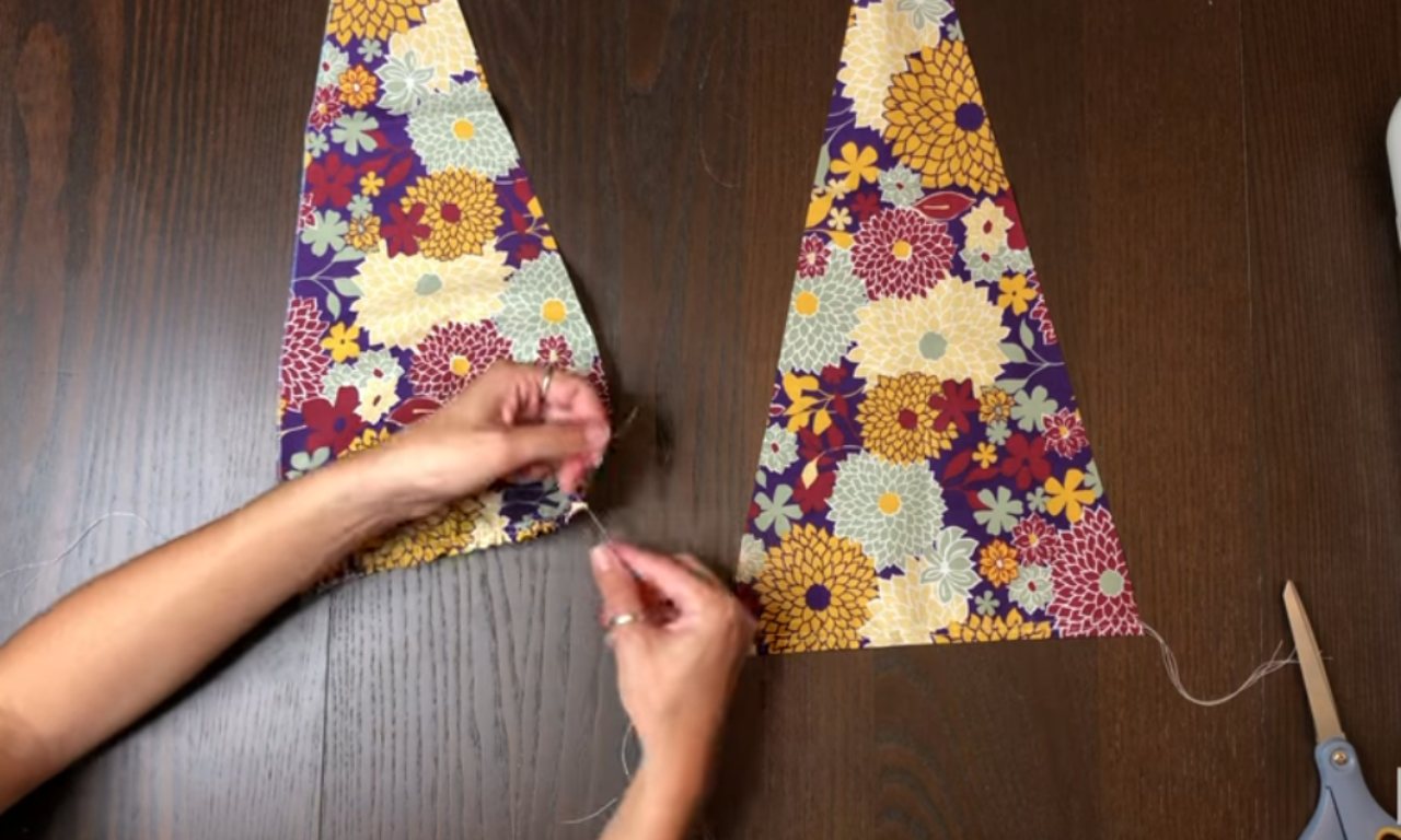 Two triangular pieces of floral fabric laid on a wooden table, with hands threading a needle to prepare for sewing.