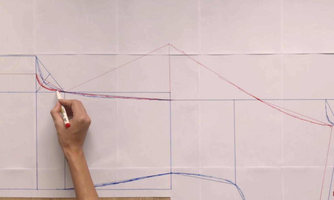 A hand using a red marker to draw curved lines on a paper sewing pattern for a wrap jumpsuit.