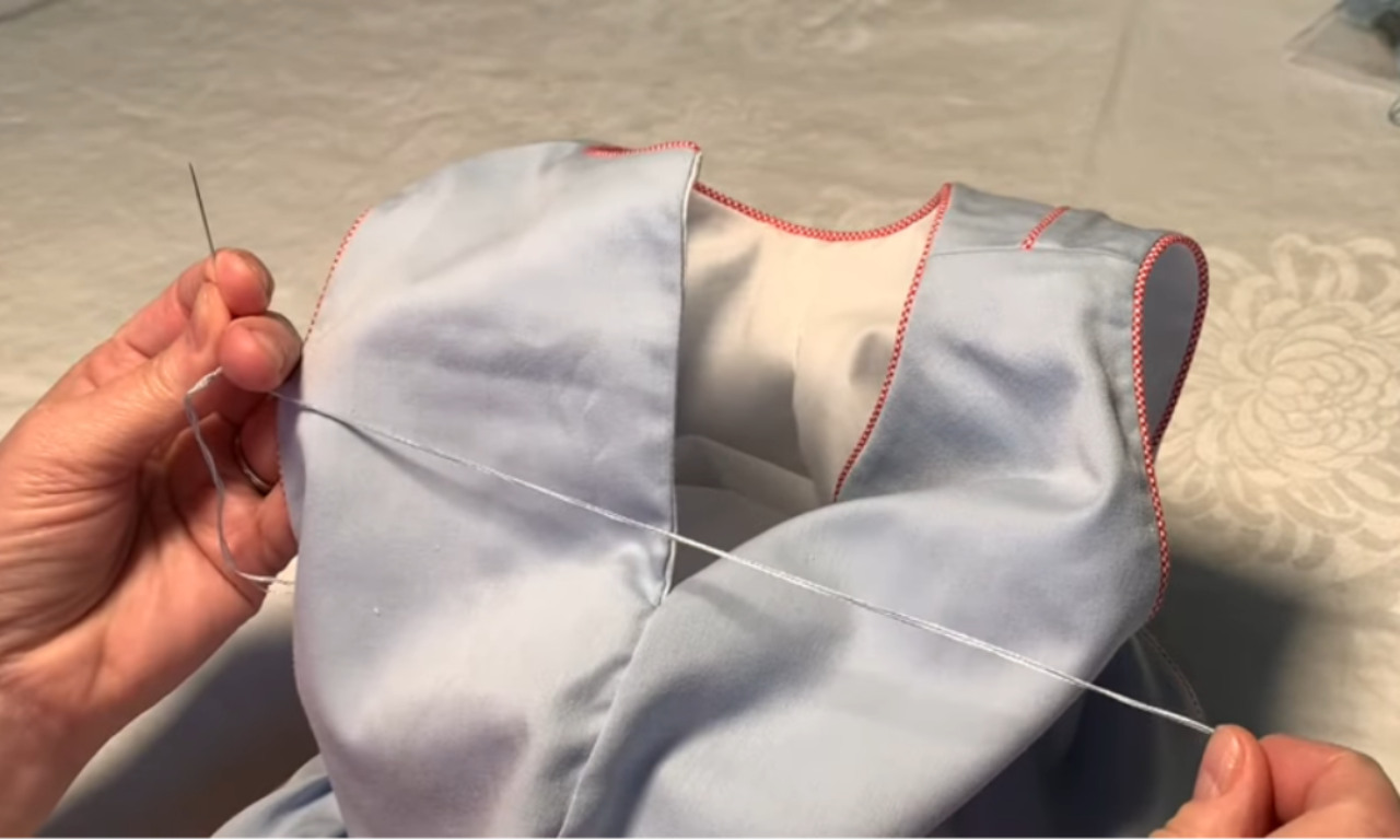 A sewer's hands creating the initial guide loops with a needle and light blue thread, the first step in how to sew a button loop by hand on a dress.