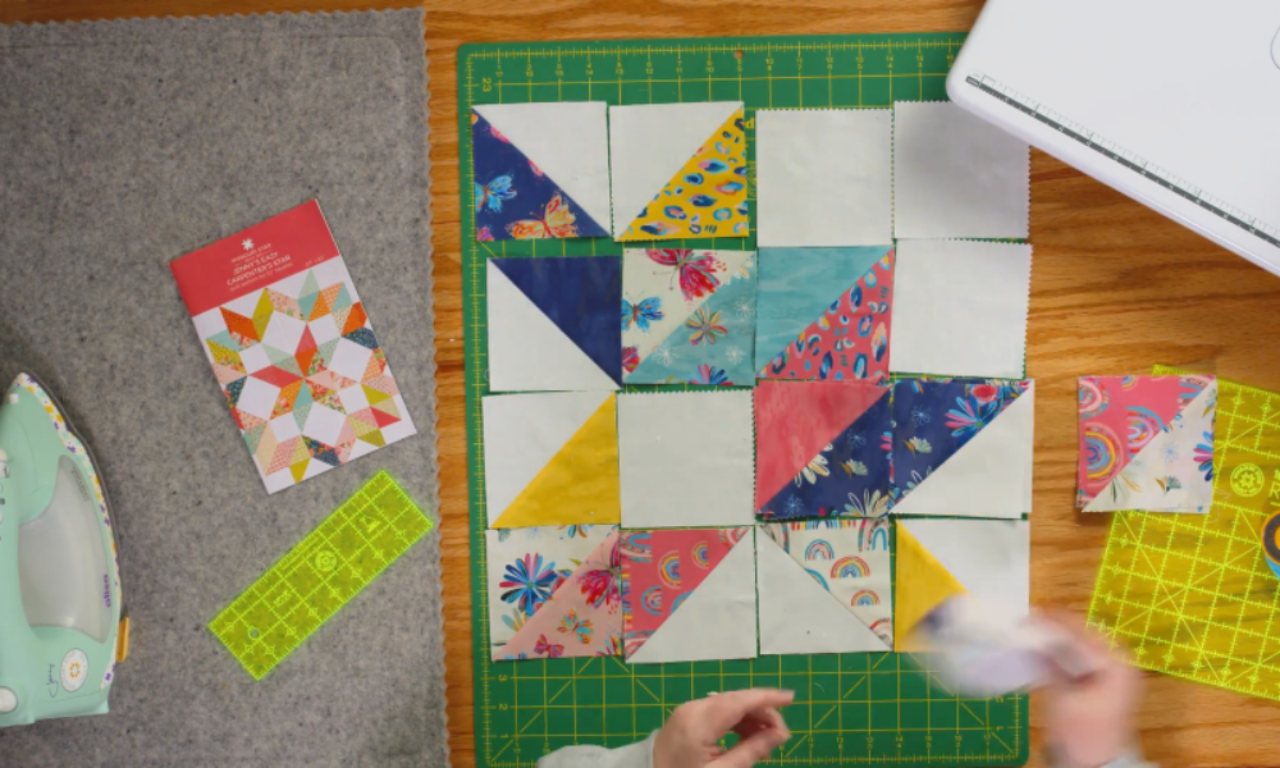 A work-in-progress quilt layout on a green cutting mat, showing small half-square triangle blocks arranged into a larger Carpenter’s Star pattern. Bright fabrics with rainbow, floral, and geometric prints are mixed with solid white pieces. Tools like a quilting ruler, rotary cutter, and an iron sit nearby.