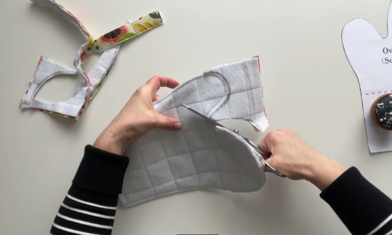 A sewer using scissors to cut along a traced pattern line on a quilted fabric sandwich to create an oven mitt.