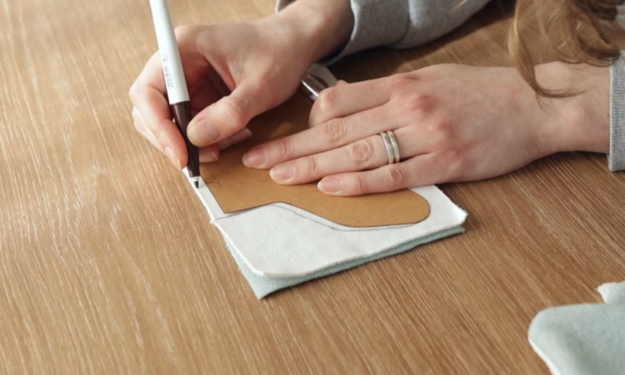 A person traces a cardboard sewing pattern for a whale fin onto two layers of fabric using a brown marker on a wooden table.