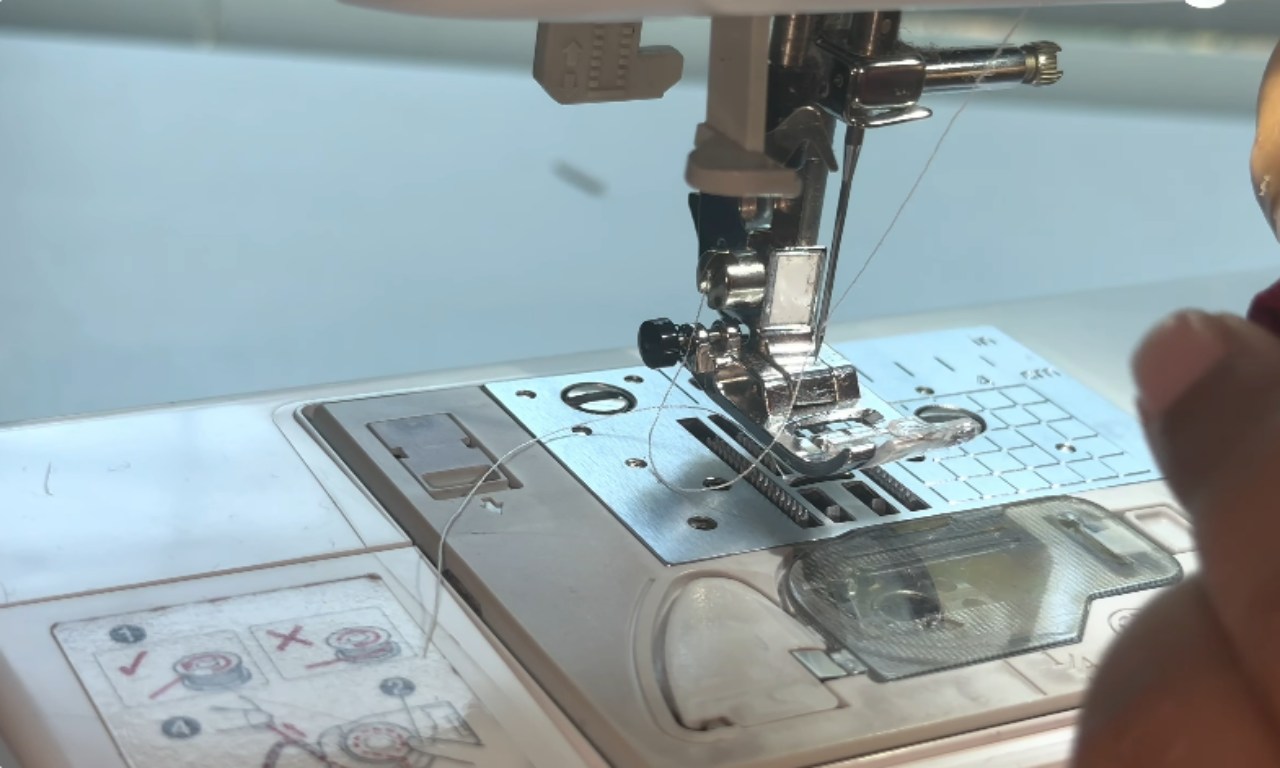 A close-up view of a sewing machine needle and presser foot, with thread visible and a person’s hand guiding the process of sewing.