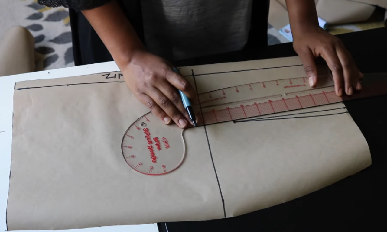 A person using a clear sewing ruler and a marker to draw a straight line on a brown paper pattern, demonstrating how to draft a maxi skirt.