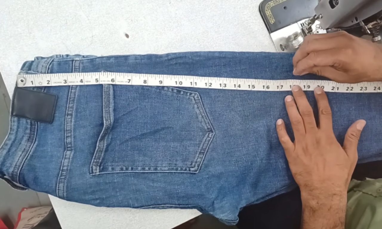 A person uses a white measuring tape to measure the desired length on the leg of a pair of blue jeans before cutting them into shorts.