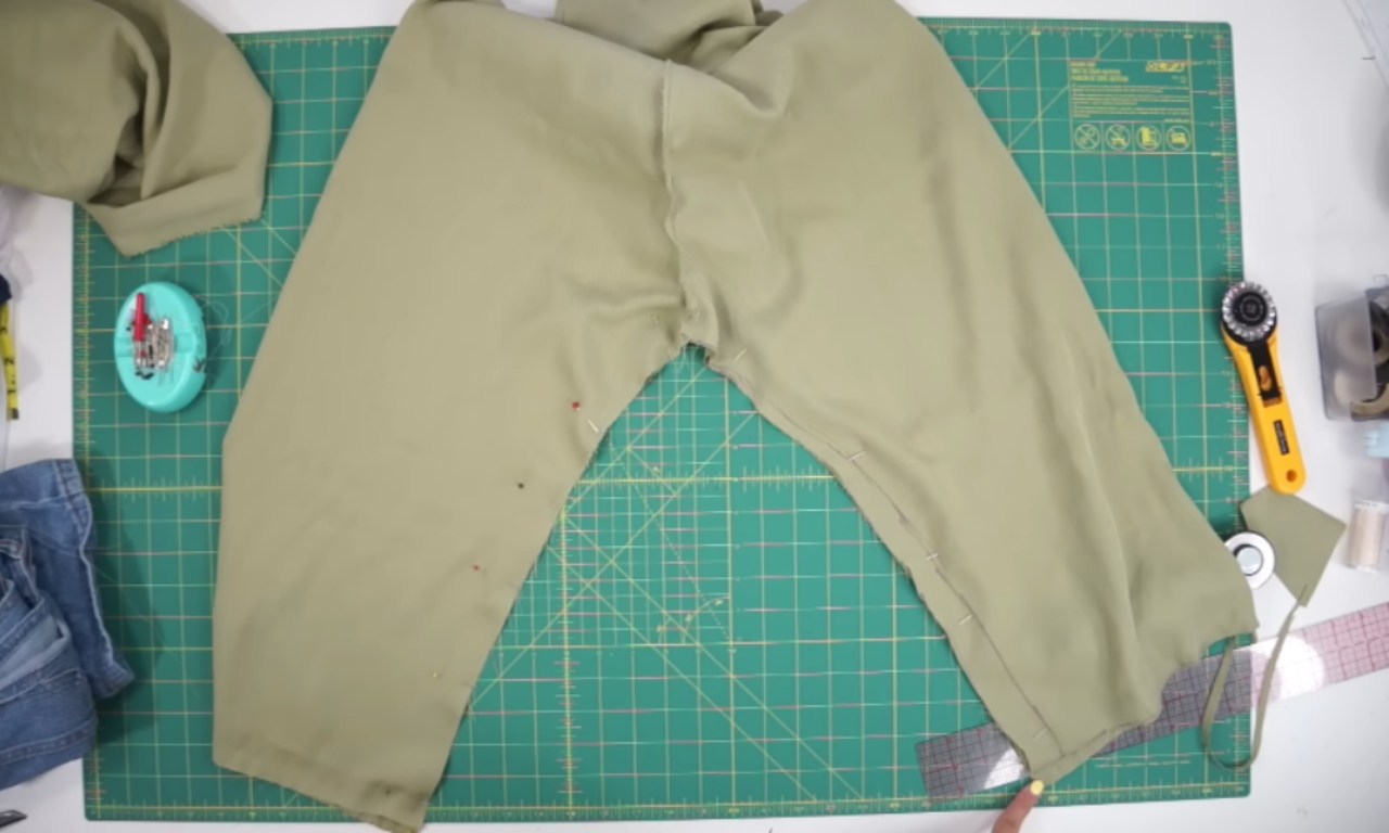A flat-lay image of green fabric laid out on a cutting mat with pins holding the fabric in place. A pair of scissors, a rotary cutter, and measuring tools are visible, with fabric edges being prepared for sewing.