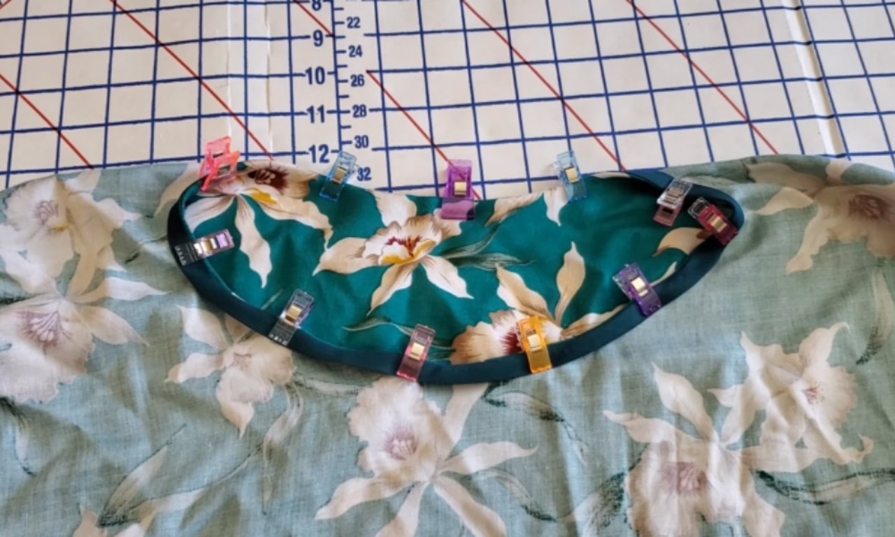 A close-up of the neckline of a floral caftan dress on a cutting mat, showing how to attach a contrasting teal facing using colorful sewing clips.