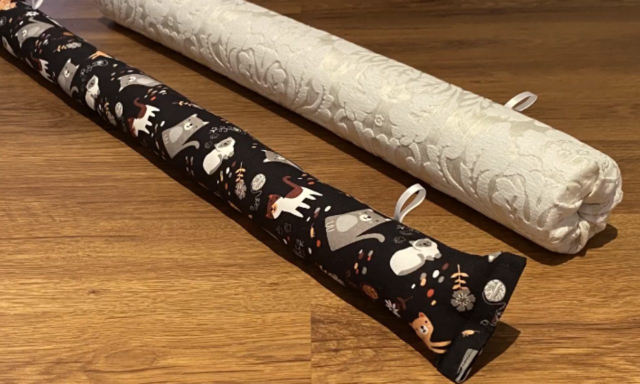 Two cylindrical fabric draft stoppers on a wooden floor, one covered in white textured fabric and the other in black fabric with a playful cat pattern.