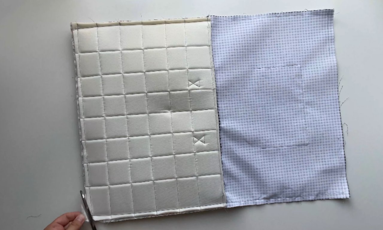 A hand using scissors to trim the edge of a piece of white quilted batting laid next to blue gingham lining fabric for a DIY laptop sleeve project.