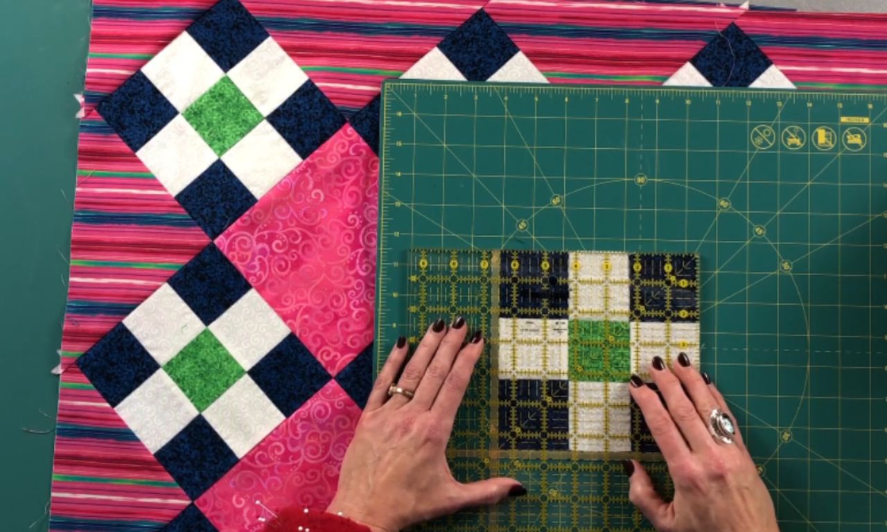 A quilter uses a square ruler to measure a finished nine-patch quilt block on a green cutting mat, with the on-point quilt top visible in the background.