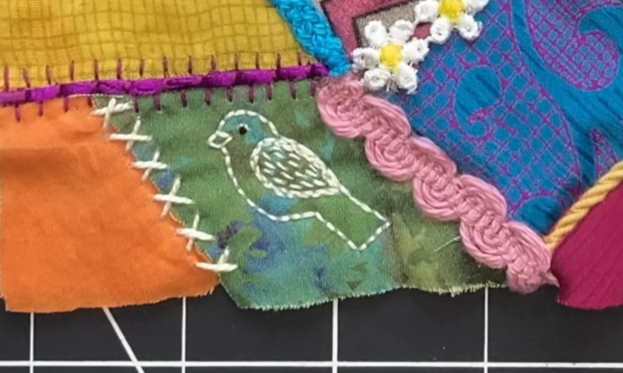 A close-up of slow stitching on a crazy quilt, featuring a small bird outlined with white embroidery floss on a green fabric patch next to pink braided trim.