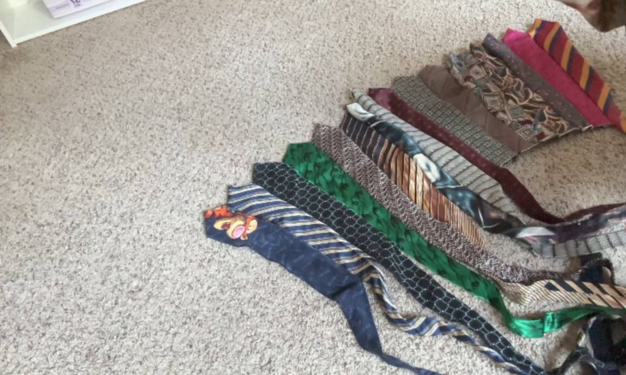 Several neckties sewn together are laid flat on a carpet, forming a circular skirt shape. The ties feature a variety of colors and patterns, and an elastic waistband is being prepared for attachment.