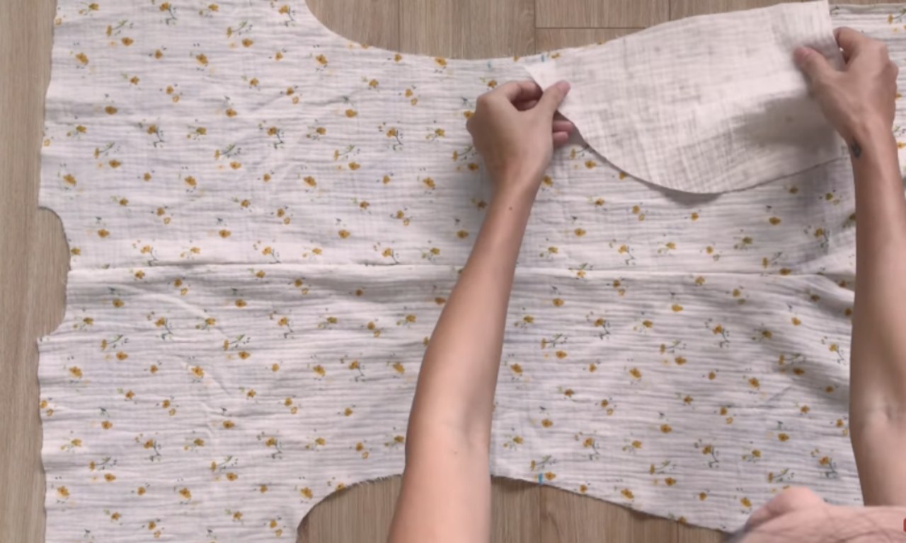 Hands arranging a pocket piece on floral fabric laid flat, preparing to attach it to a jumpsuit pattern.
