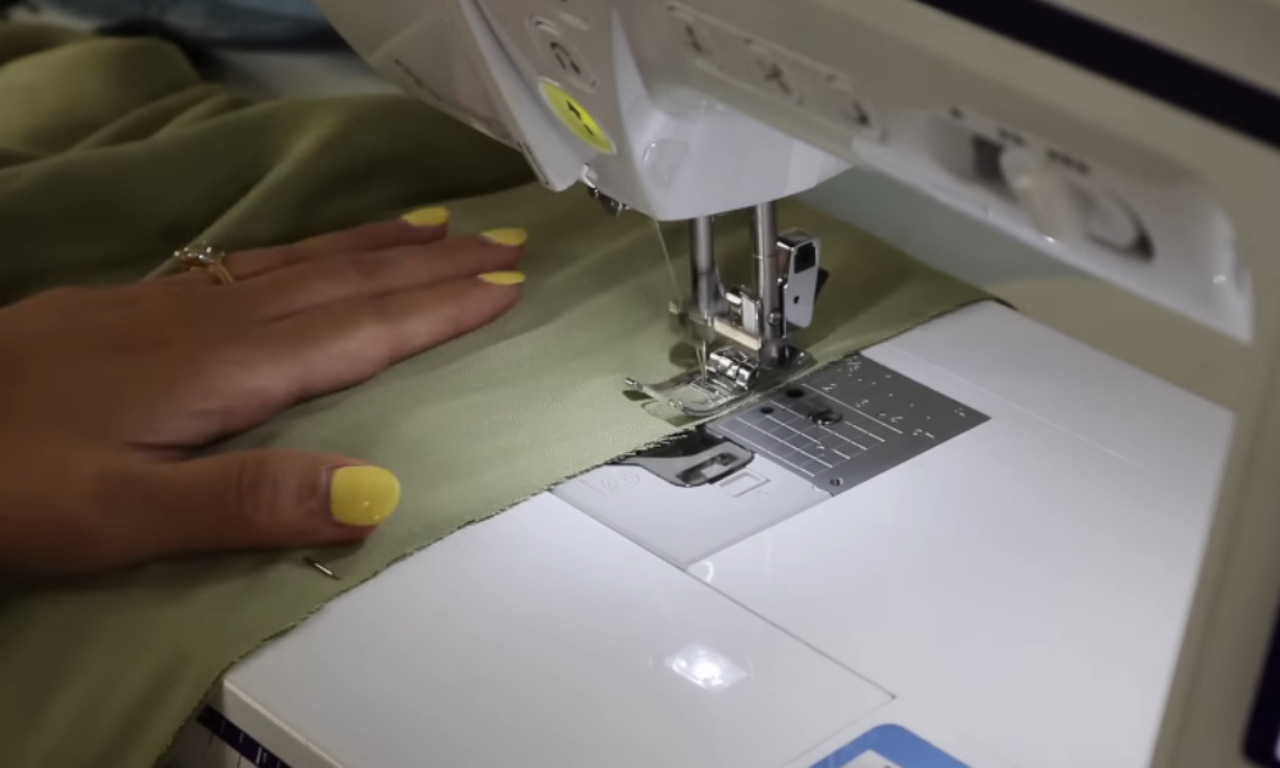 Close-up shot of a sewing machine with a person’s hand guiding green fabric under the needle. The sewing machine is stitching the fabric, and the hand is adorned with yellow nail polish.
