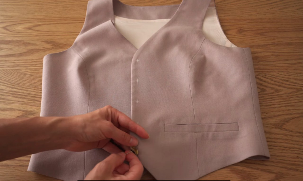 Close-up of a person's hands hand-sewing a button onto the front of a light purple waistcoat laid flat on a table.
