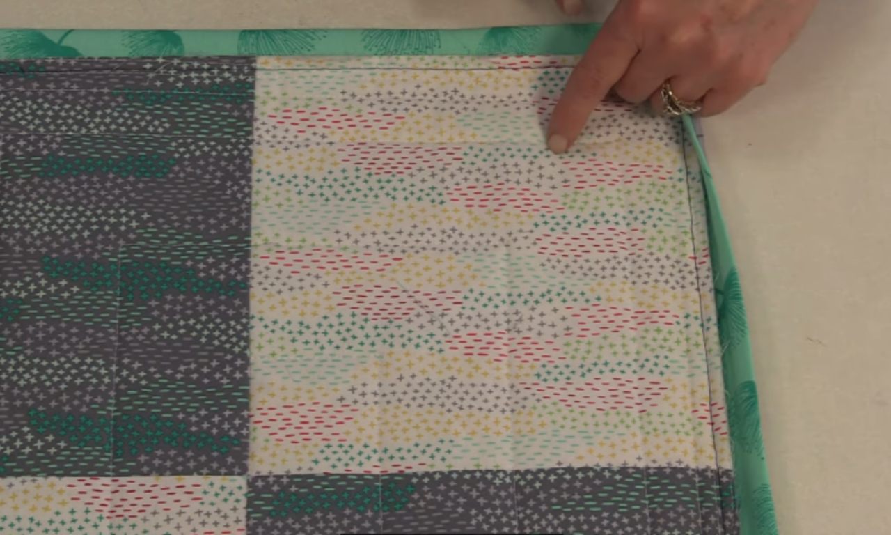 A finger points to the stitch line on the back of a quilt where the faux flanged binding has been attached, ready to be folded over to the front.