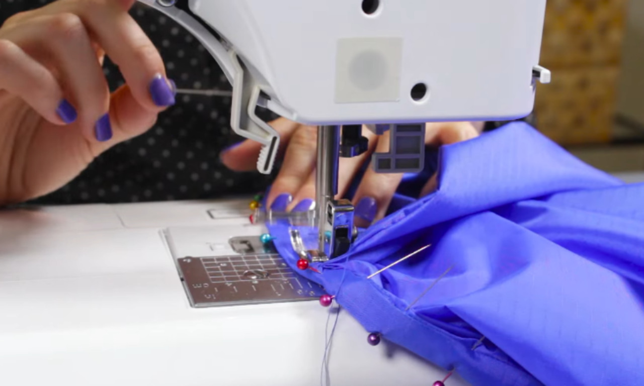 Close-up of a person’s hand sewing a bright blue fabric on a sewing machine. The sewing machine is threaded, and colorful pins hold the fabric in place. The person’s nails are painted purple.