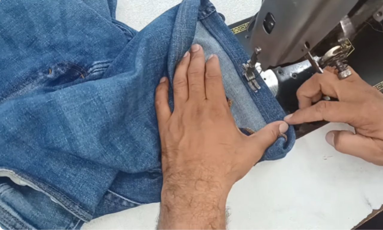 A sewer's hands carefully guide the folded hem of a blue denim short under a sewing machine to stitch it in place.
