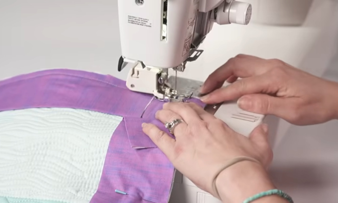 Close-up of a person sewing purple fabric onto a quilt using a sewing machine, carefully guiding the material with their hands.