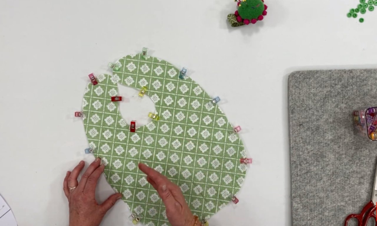 A piece of green patterned fabric cut in the shape of a baby bib, held in place with colorful sewing clips around the edges. A person’s hands are adjusting the fabric on a white table with sewing tools nearby.