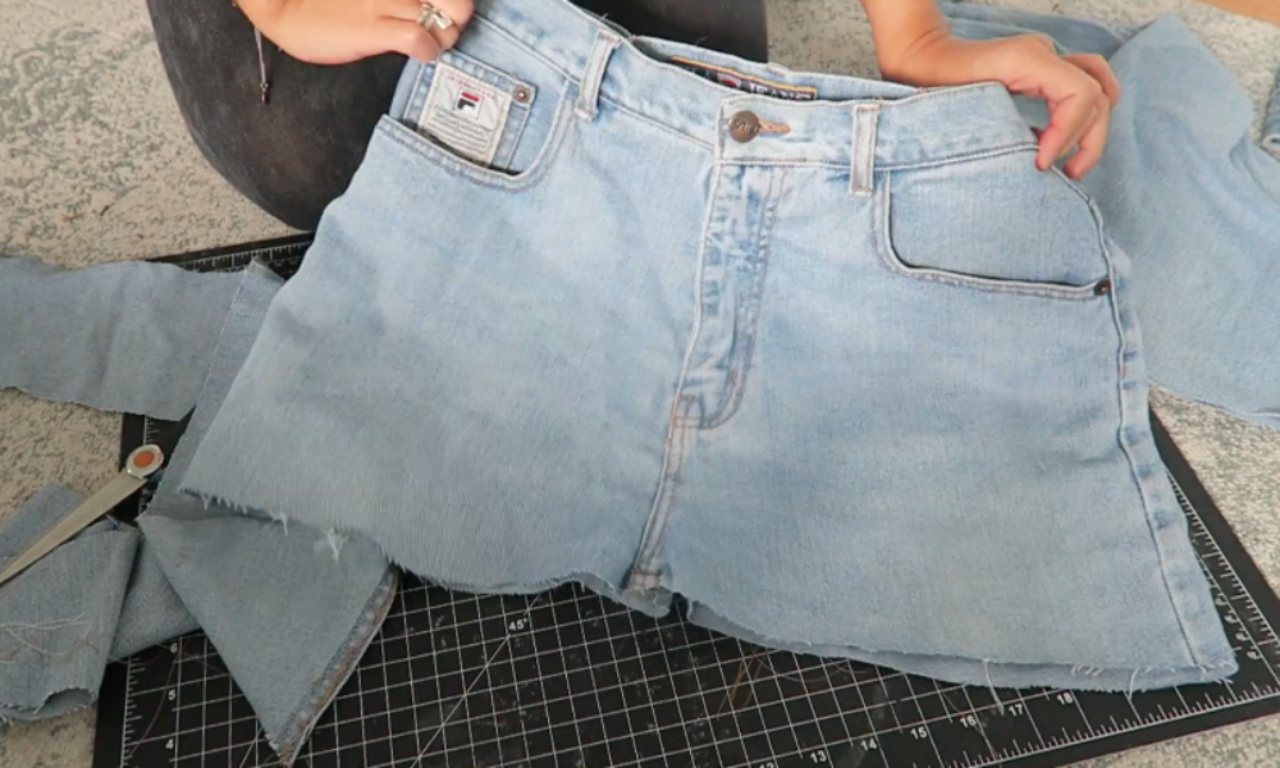 Progress on a DIY denim skirt, showing light-wash jeans on a cutting mat with the legs cut off and the inseam opened up, creating fabric flaps ready to be made into a skirt.