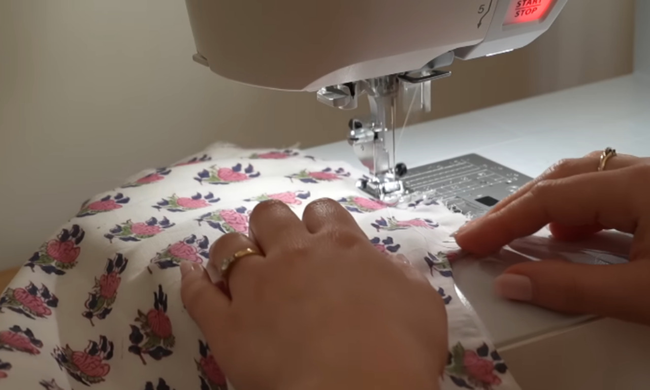 Close-up of hands guiding floral fabric through a sewing machine, stitching the material. The fabric is white with a pink and purple floral print.