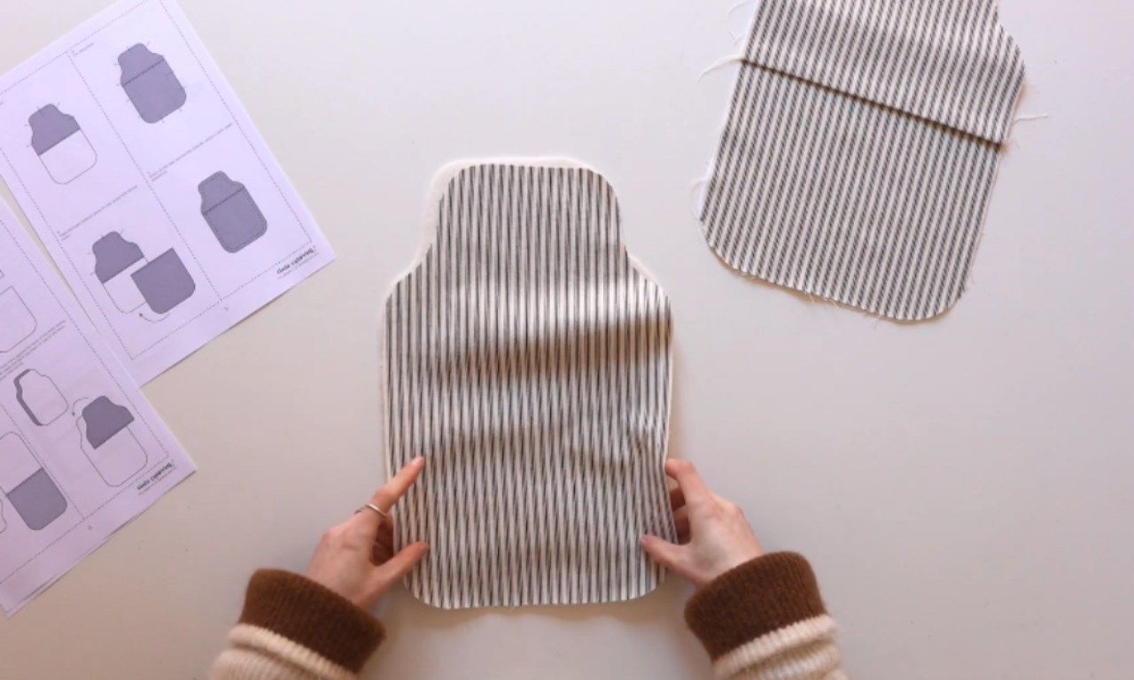 Hands holding a cut piece of striped fabric shaped like a hot water bottle, with another fabric piece and printed sewing instructions visible on the table.
