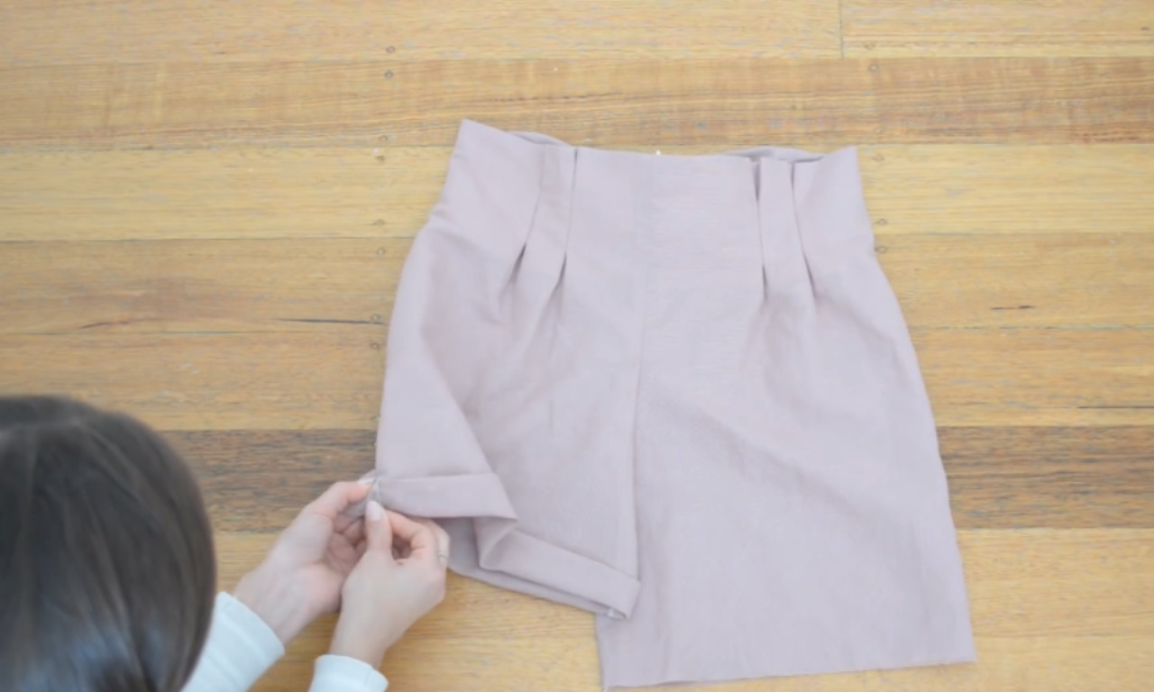 A person pins the cuff of a pair of light pink paper bag shorts on a wooden surface, preparing to sew the hem.