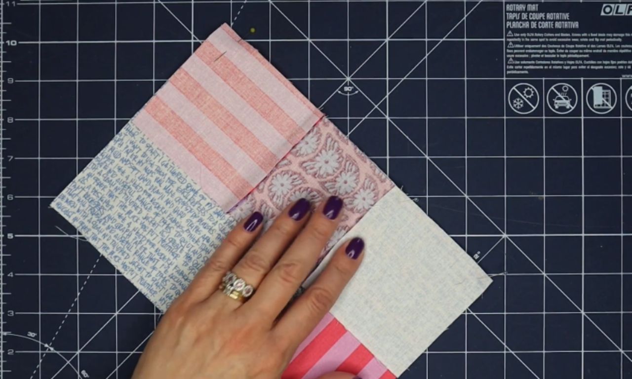 A hand with purple nail polish holds together four rectangular fabric pieces sewn into a block, laid out on a black cutting mat with a white grid. The fabrics have different patterns including stripes, floral, and script text.