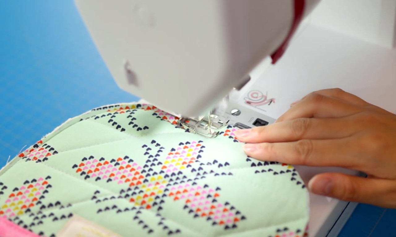 A close-up view of a sewing machine stitching the pink binding onto a light green quilted pot holder with a geometric pattern.