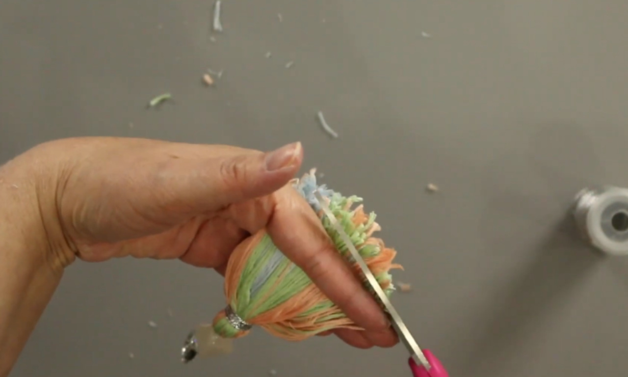 A crafter's hand holds a colorful DIY yarn tassel keychain while using scissors to trim the bottom for a clean, finished edge.