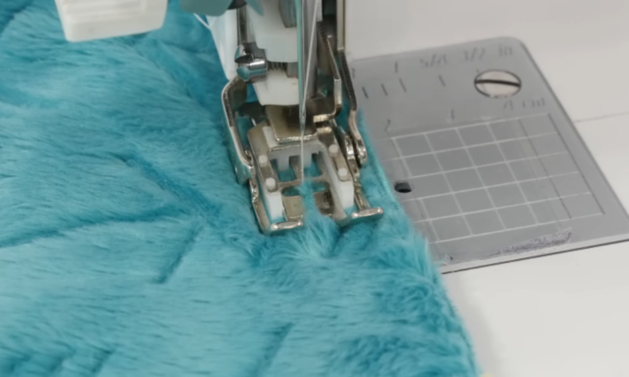 A close-up view of a sewing machine stitching through blue minky fabric with an arrow pattern. The machine uses a walking foot to guide the thick, plush fabric evenly.