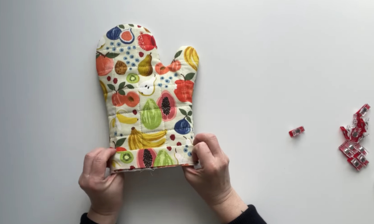 A person's hands finishing the binding on a homemade quilted oven mitt with a colorful fruit pattern.