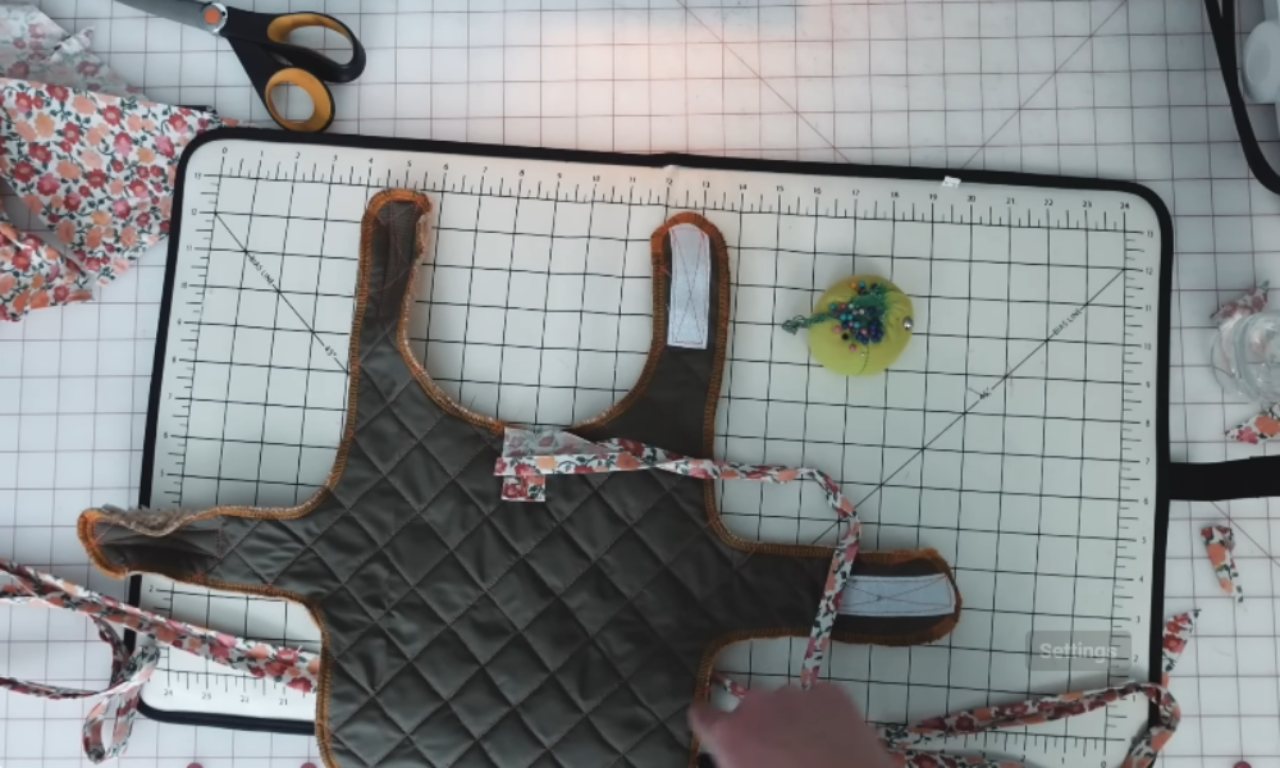A quilted dog jacket pattern with floral straps laid flat on a cutting mat, alongside scissors, fabric scraps, and a pincushion.