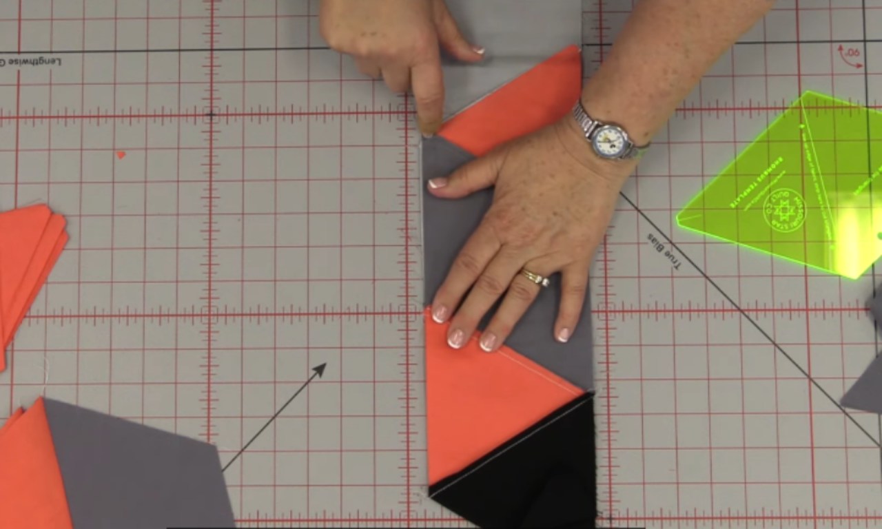 A close-up of a person arranging fabric triangles in shades of coral, gray, and black on a gridded cutting mat. The hands are positioning the pieces to form a geometric block pattern, likely for a tumbling block quilt design.