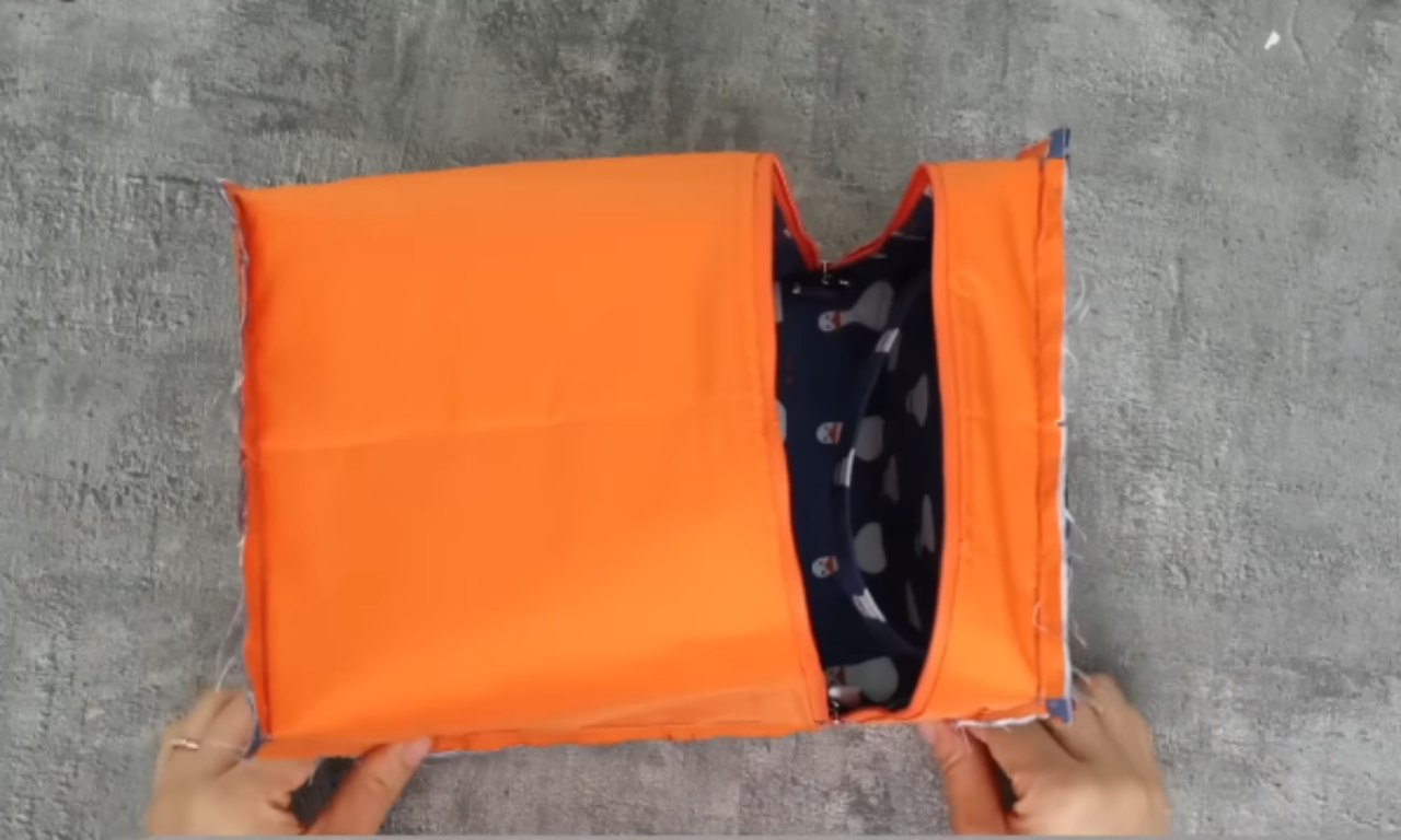 A close-up view of the inside of the insulated lunch bag, showing bright orange inner lining with a peek of the blue duck-patterned fabric. Hands are holding the bag open on a gray surface.