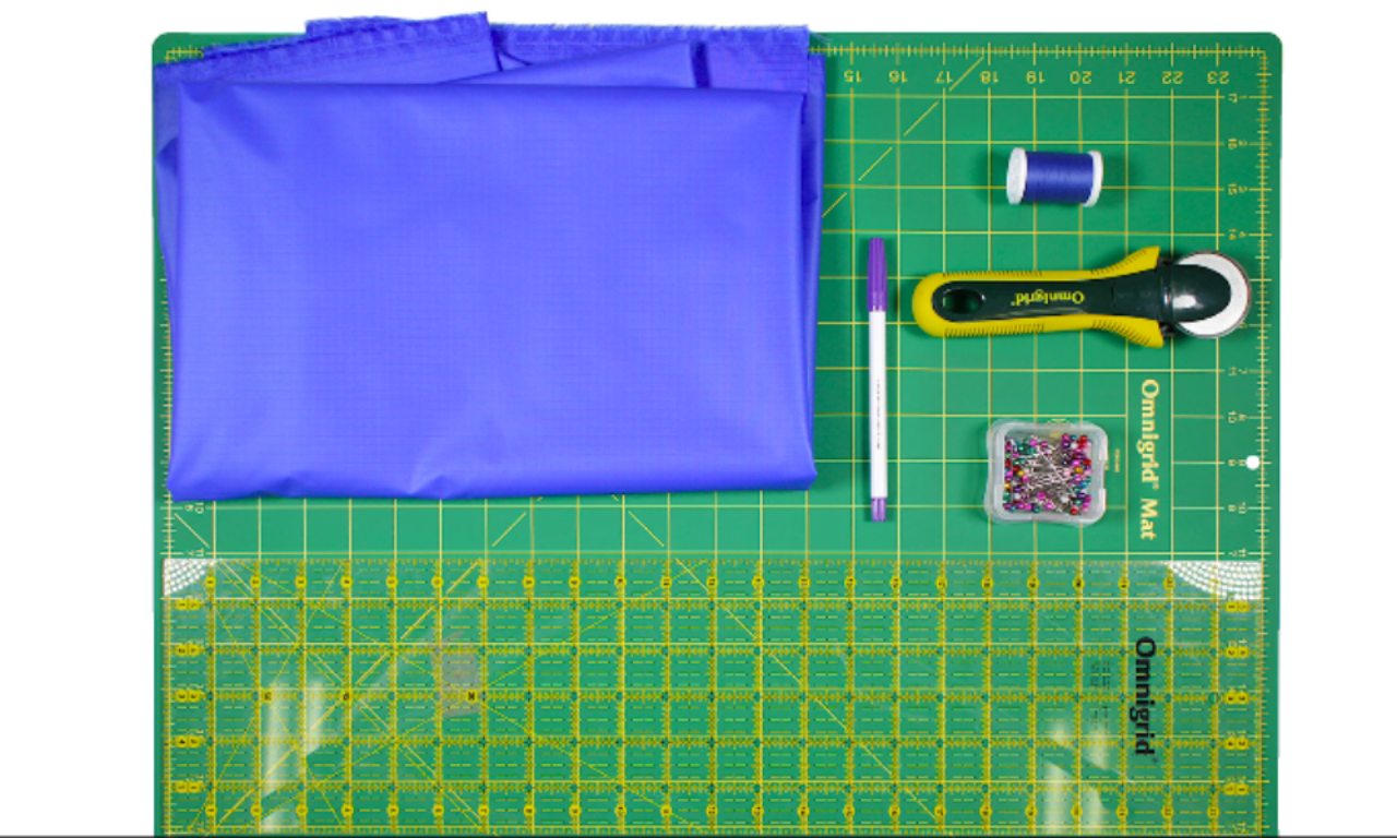 A flat lay of materials for making a rain poncho. The items include folded blue fabric, a white fabric marker, a rotary cutter, a container of colorful pins, and a yellow grid ruler on a green cutting mat. The cutting mat has measurement lines for precise cutting.