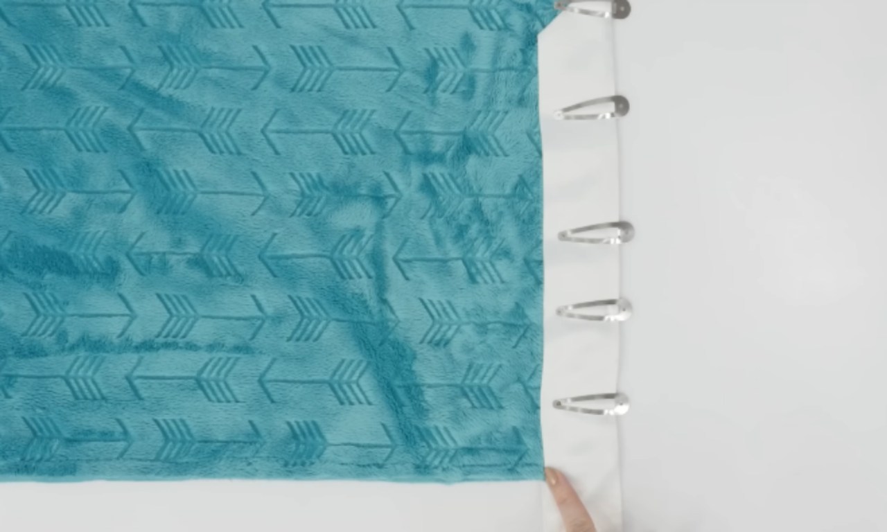 A top-down image showing blue minky fabric pinned to a white satin border using large safety pins, preparing the edges for stitching.