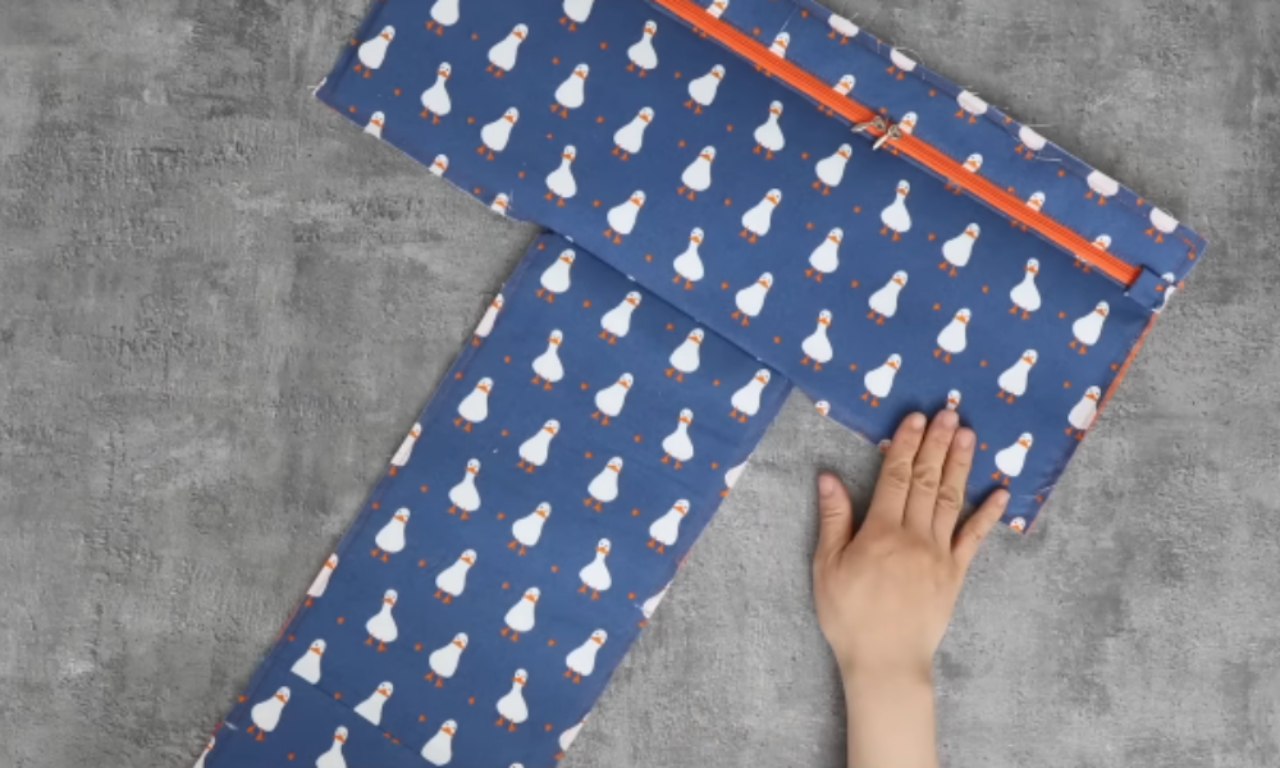 A flat, partially sewn piece of blue fabric with a duck pattern and an orange zipper is laid out on a gray surface. A hand is placed on the fabric.