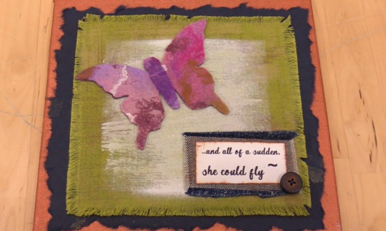 A handmade mixed-media canvas featuring a large pink and purple felt butterfly. The background is layered with orange, dark blue, and green fabric. In the bottom corner, a denim patch displays the quote, "...and all of a sudden, she could fly."