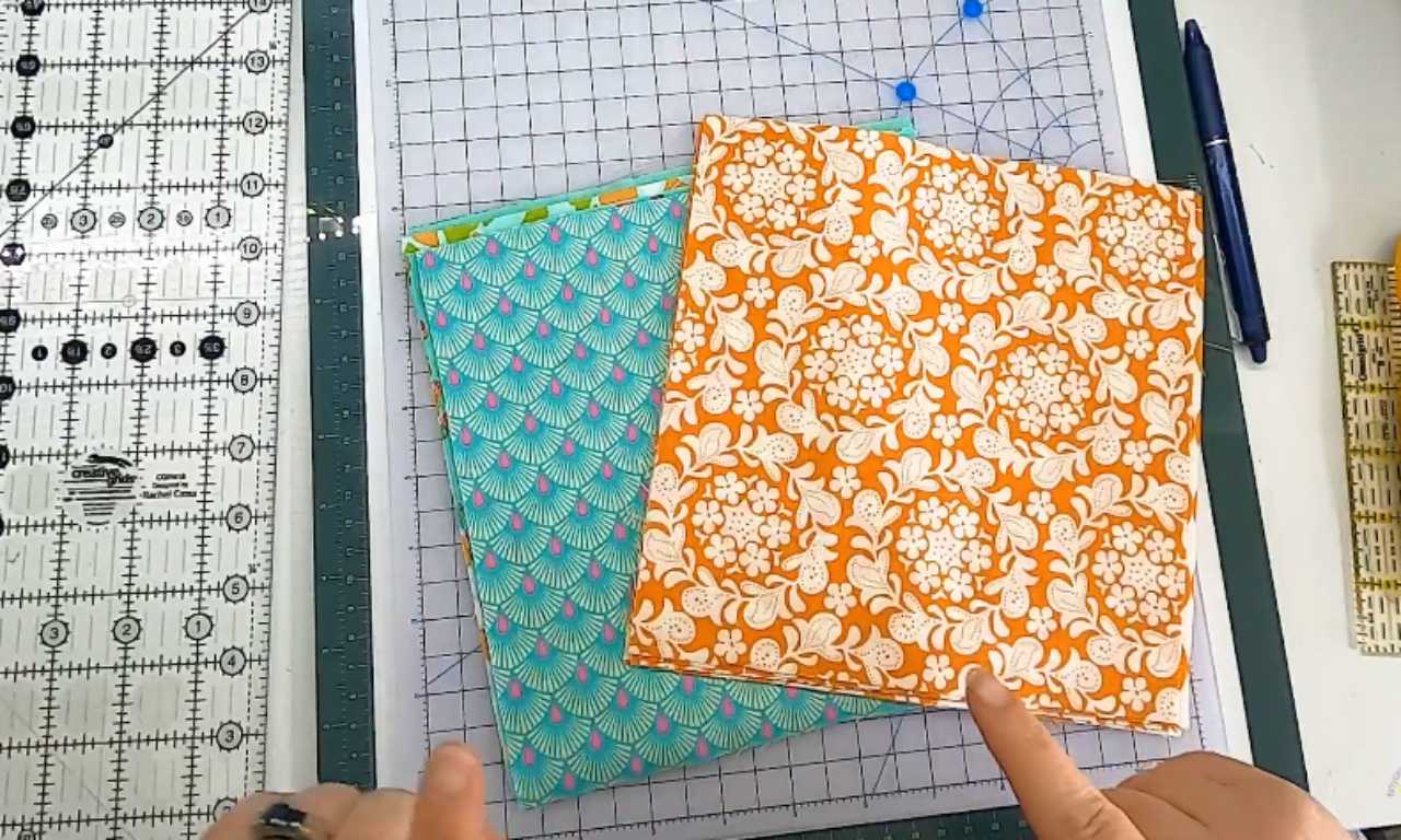 A quilter's hands showing two stacks of patterned fat quarter fabrics in orange and aqua, laid out on a white cutting mat.