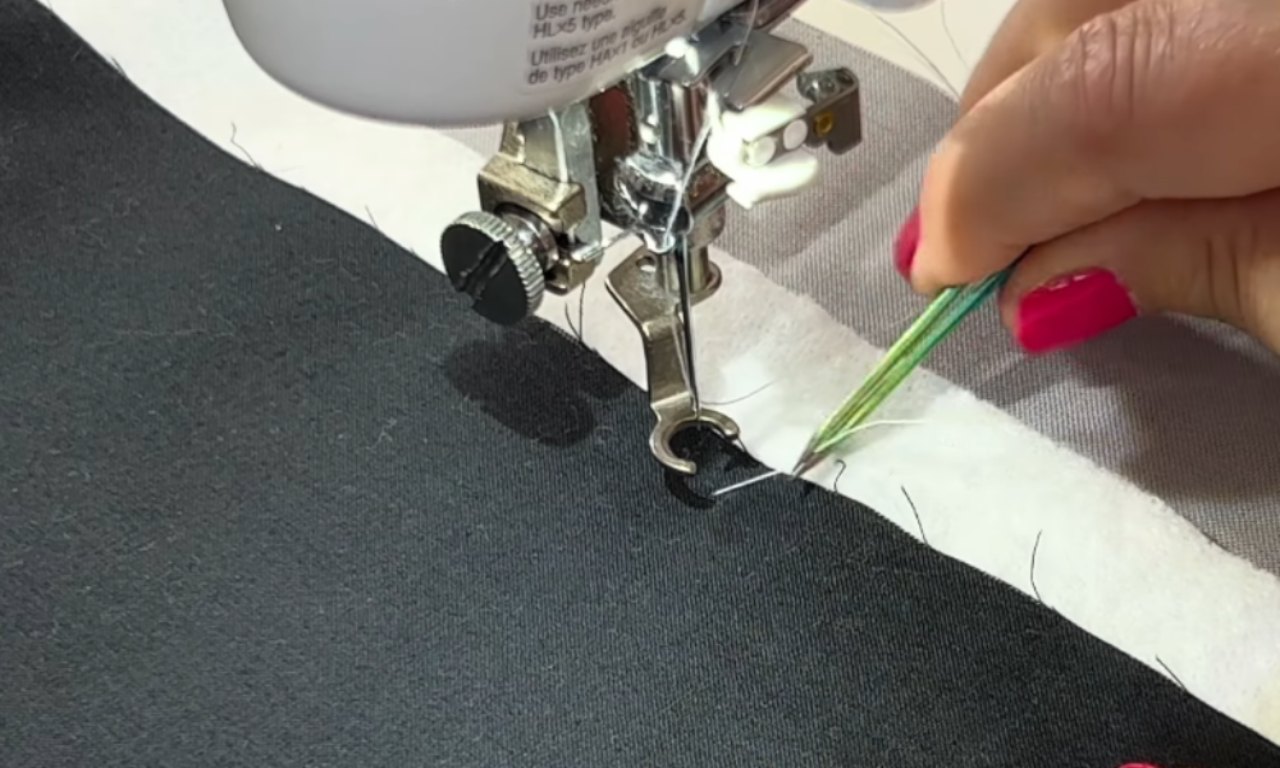 Hand with pink-painted nails using tweezers to guide thread under a sewing machine needle on black and white layered fabric.