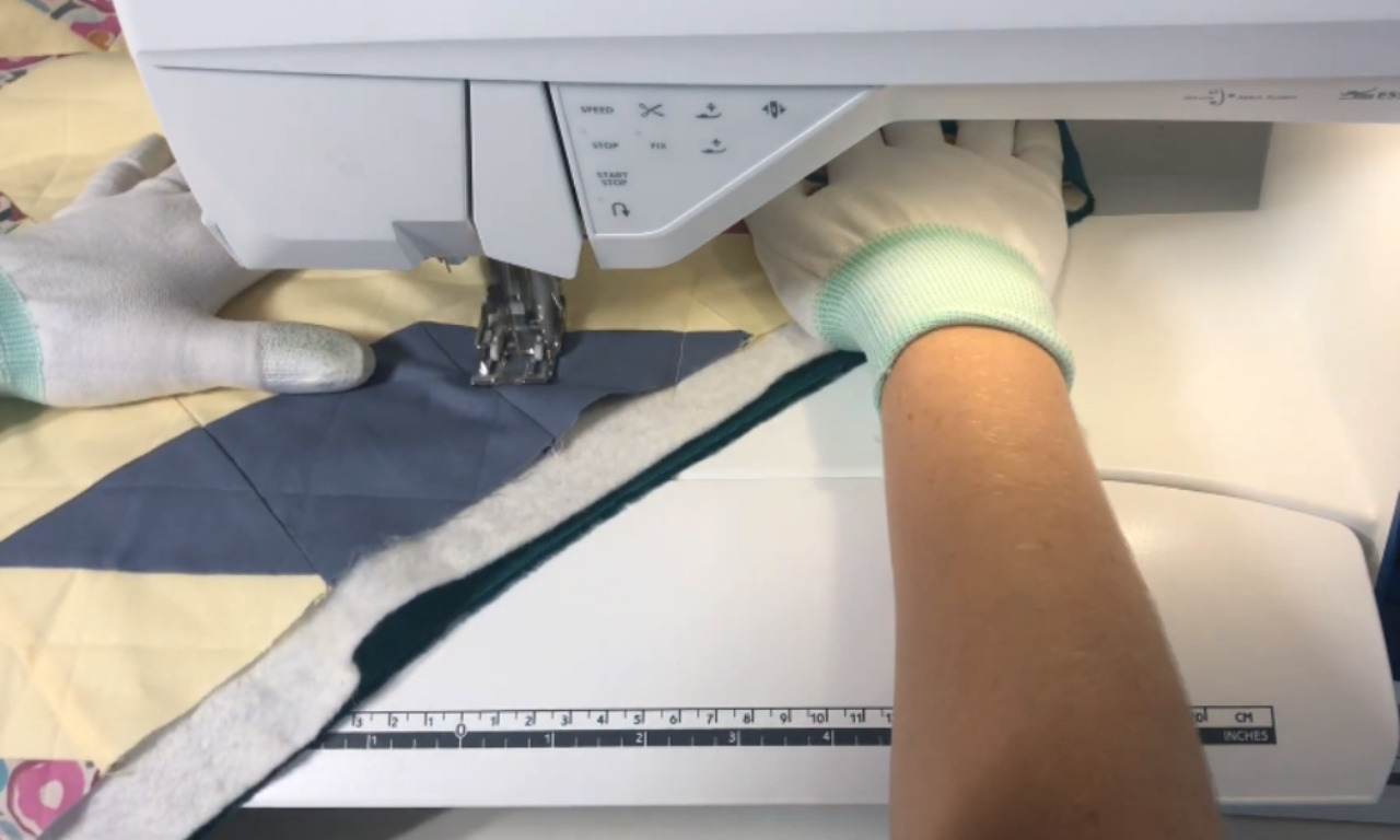 Person quilting on a sewing machine, wearing white grip gloves, guiding a layered quilt sandwich under the walking foot.