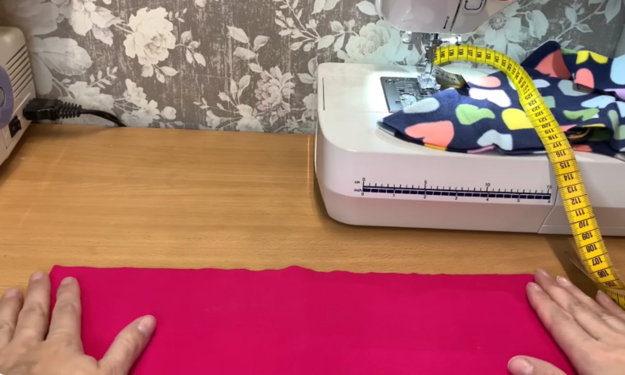 A person prepares a piece of bright pink fabric for sewing on a wooden table, with a sewing machine and measuring tape in the background.