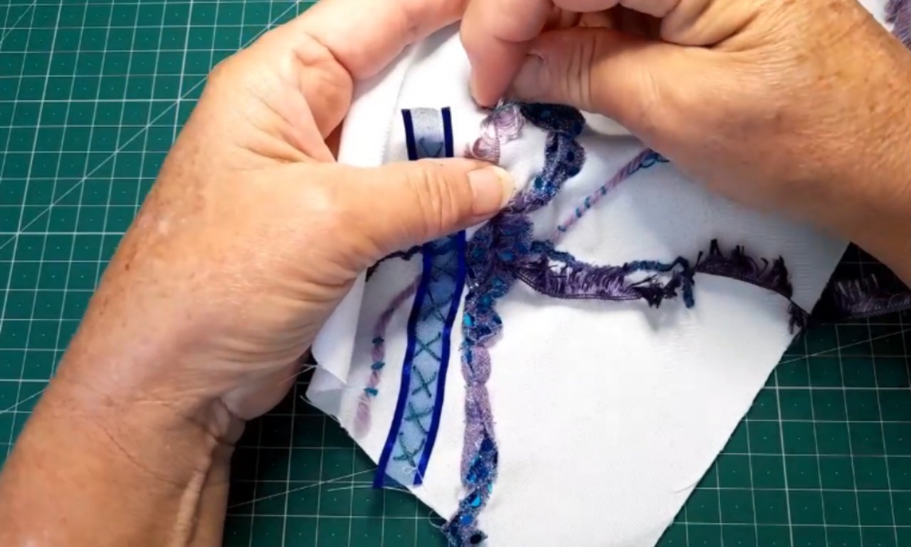 A close-up of hands demonstrating the couching stitch, using a needle to tack down a decorative purple and blue yarn onto a piece of white fabric.
