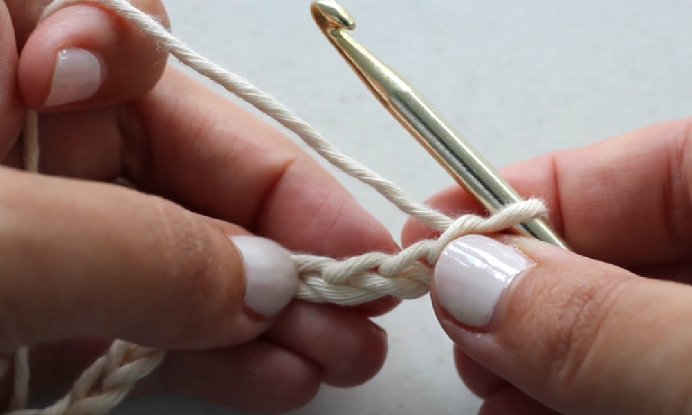 A close-up of hands holding a gold crochet hook and off-white yarn to demonstrate how to make a crochet foundation chain stitch.