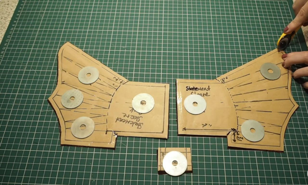Paper sewing patterns for statement sleeves laid out on a green cutting mat, held in place with metal washers, while a person uses a rotary cutter.