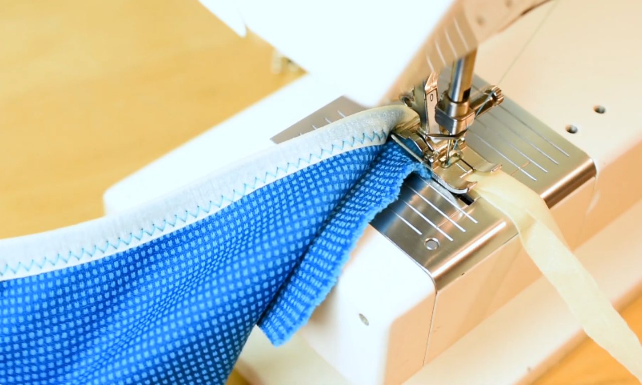 A sewing machine presser foot stitching along the edge of white fold-over elastic that is folded over the blue fabric, creating a finished seam.