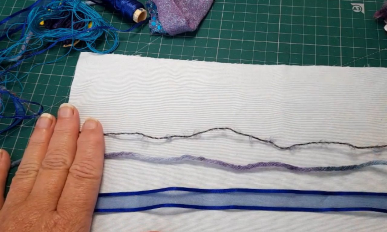 A hand holding three different trims—a dark yarn, a purple yarn, and a blue ribbon—laid out horizontally on a piece of white fabric, ready for a couching stitch project.